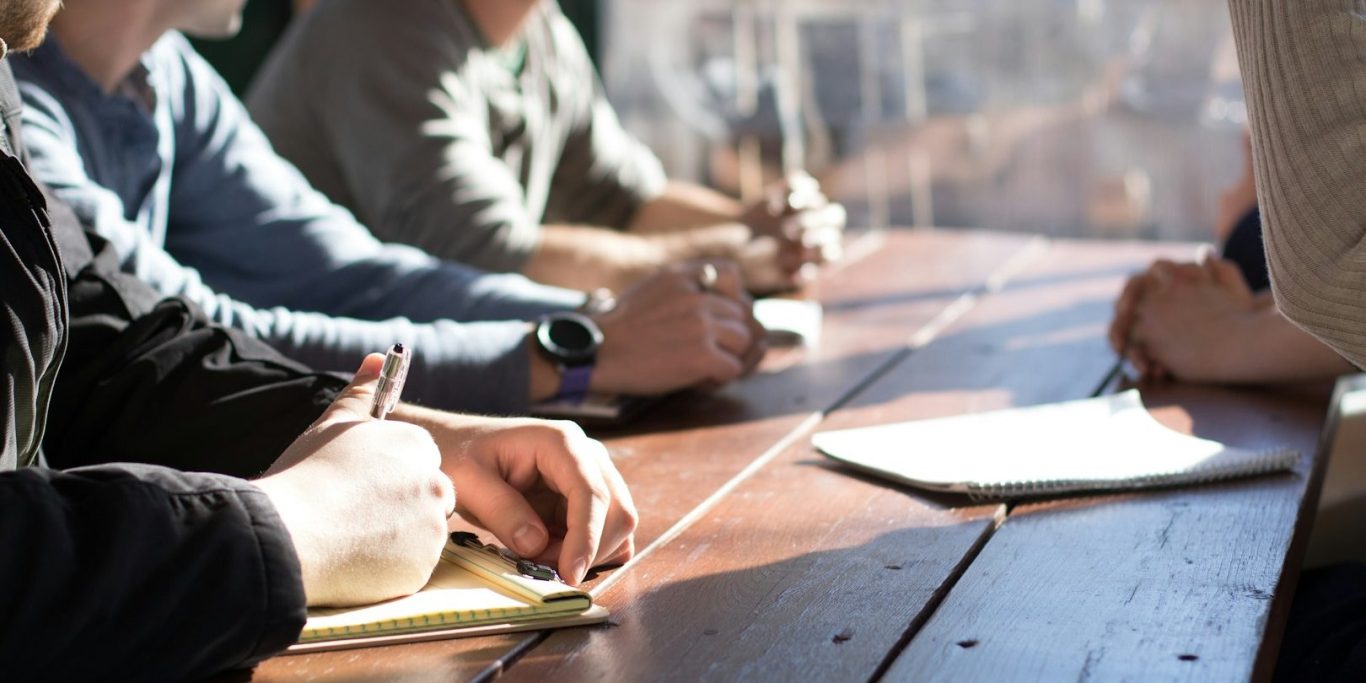 Hands of several people taking notes at a wooden table during a meeting.